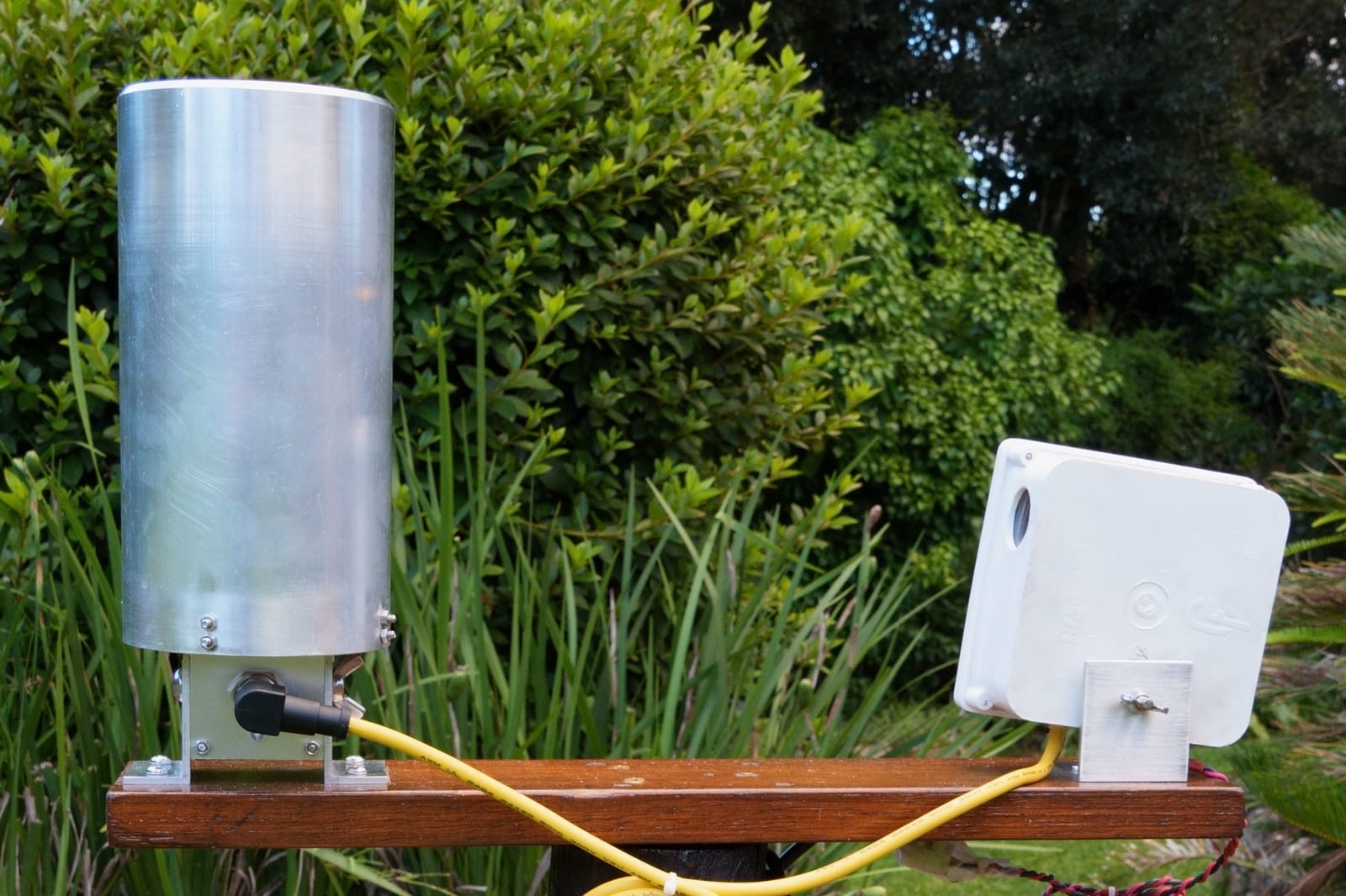 A Midge rain gauge, an aluminium tube on a timber mount, with its cable running to a white enclosure beside it.