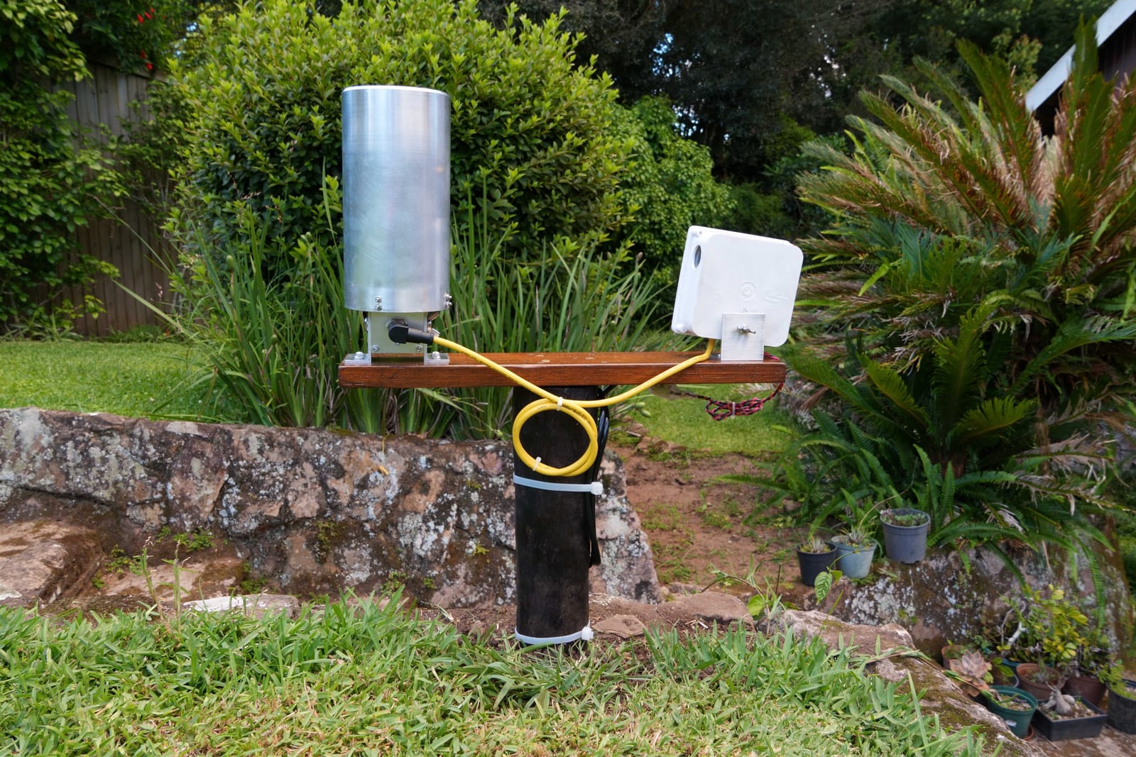 The same Midge rain gauge from further back, on a post among shrubs and a stone wall.